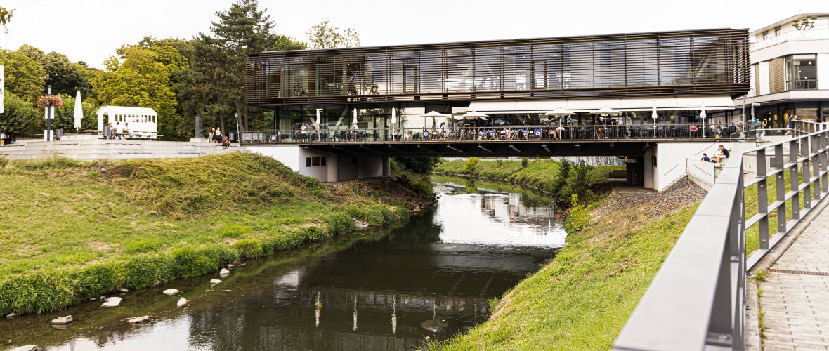 Modernes Gebäude mit Glasfassade, das eine Brücke über einen Fluss bildet, umgeben von Grünflächen und einem Gehweg mit Geländer