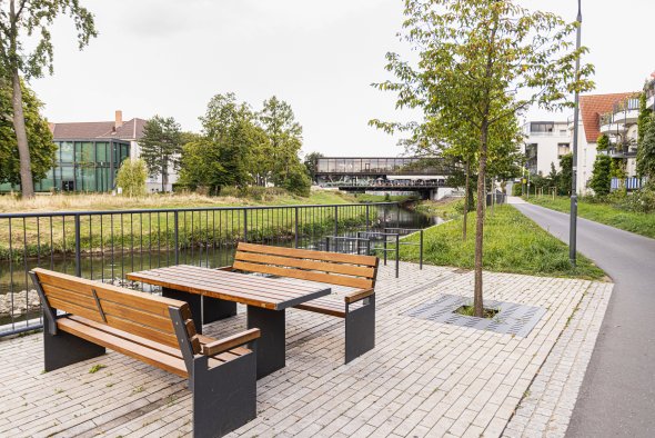 Holztisch mit zwei Bänken auf gepflastertem Platz neben einem Fluss mit Brücke und Bäumen im Hintergrund