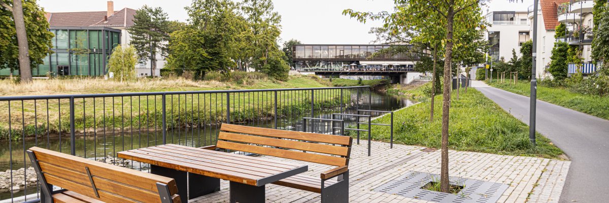 Holztisch mit zwei Bänken auf gepflastertem Platz neben einem Fluss mit Brücke und Bäumen im Hintergrund
