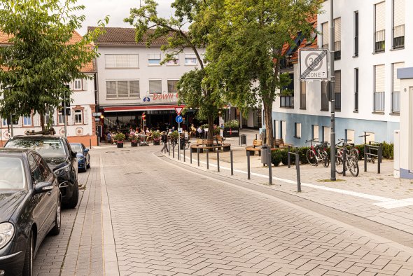 Blick in eine ruhige Straße mit geparkten Autos links, Fahrradständern rechts und einem Restaurant mit Außenbestuhlung im Hintergrund