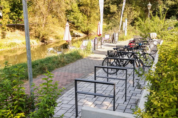 Fahrräder stehen an Fahrradständern auf gepflastertem Weg neben einem Fluss mit Sonnenschirmen und Bäumen im Hintergrund