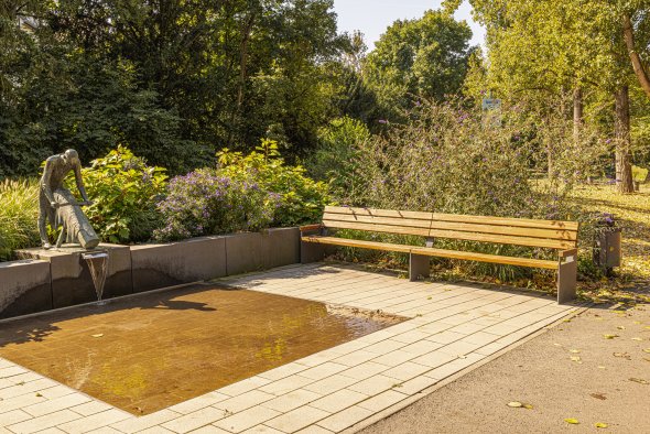 Park mit einer Holzbank neben einem flachen Wasserbecken und einer Bronzeskulptur, die Wasser in das Becken gießt
