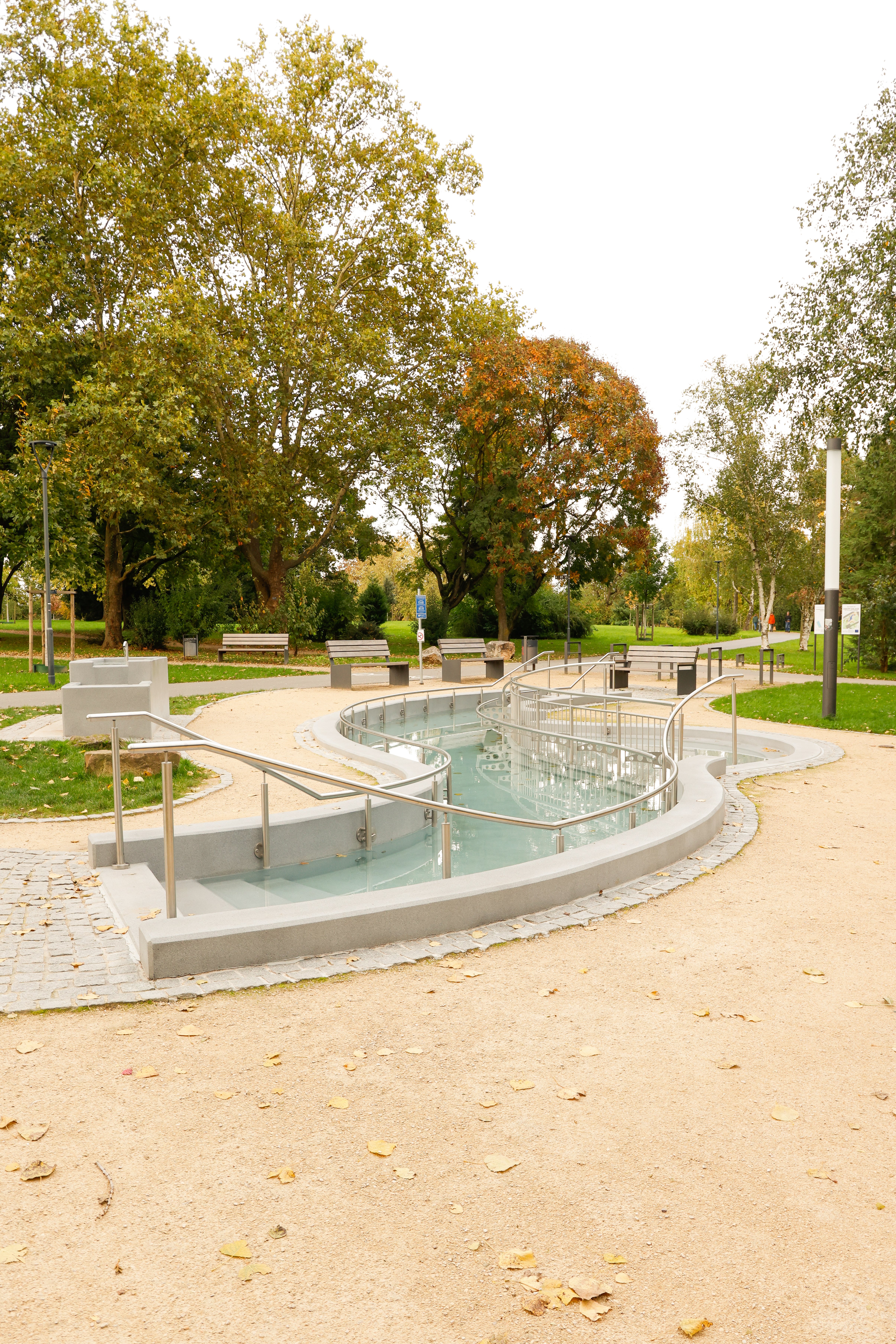 Geschwungener Wasserlauf mit Handläufen und Bänken in einem Park mit Bäumen und Wegen