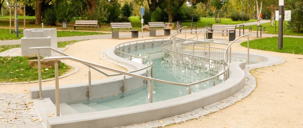 Geschwungener Wasserlauf mit Handläufen und Bänken in einem Park mit Bäumen und Wegen