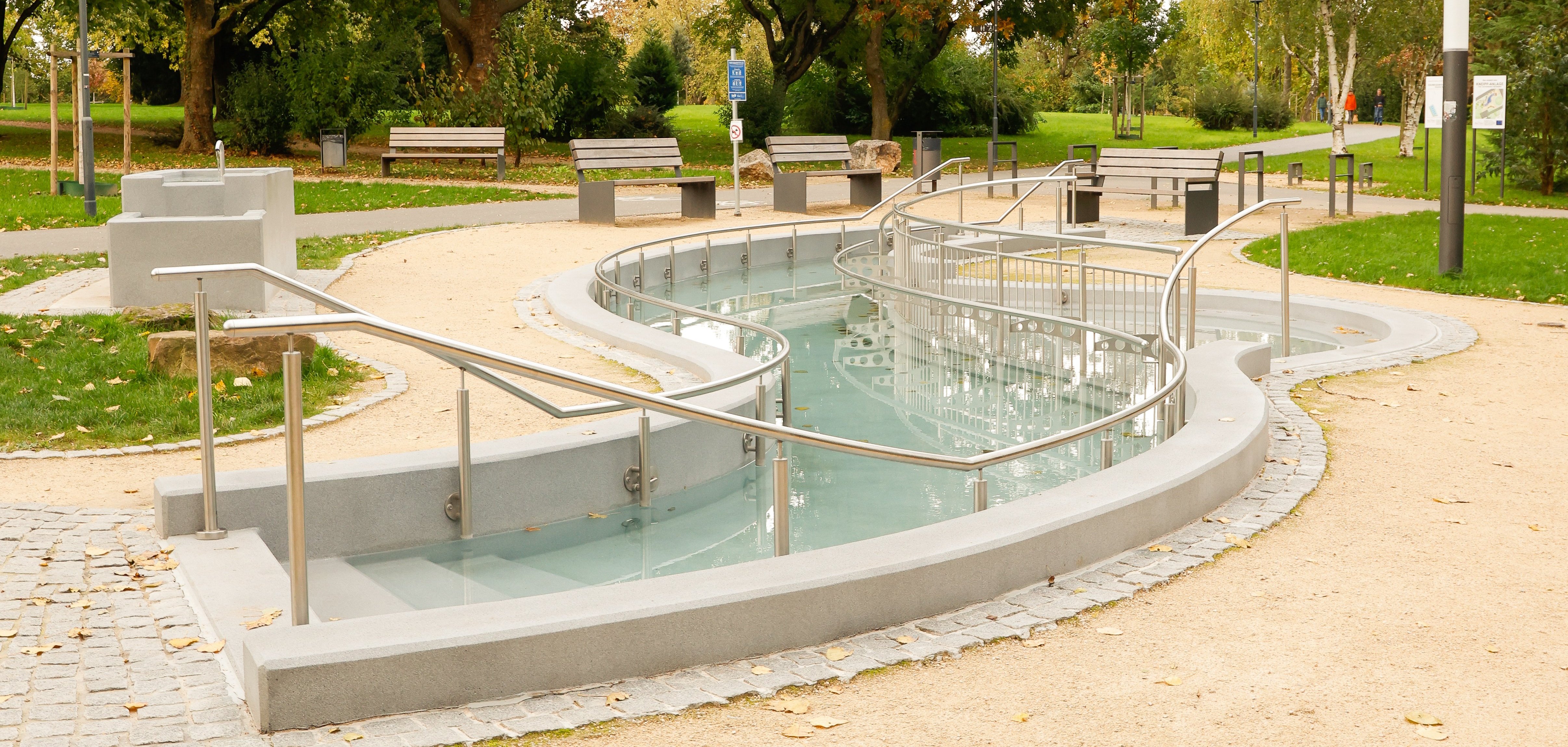 Geschwungener Wasserlauf mit Handläufen und Bänken in einem Park mit Bäumen und Wegen