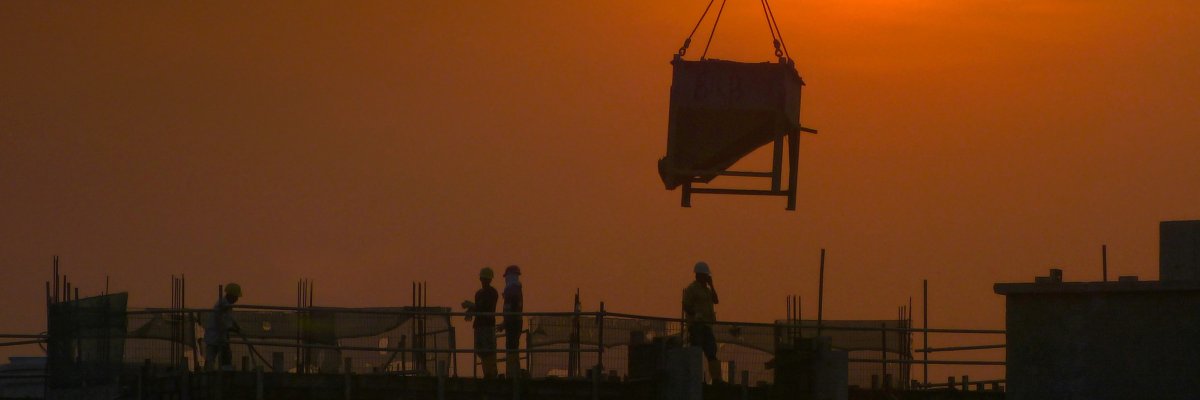 Silhouetten von Bauarbeitern auf einem Dach bei Sonnenuntergang mit einem Kranhaken, der einen Lastbehälter hebt