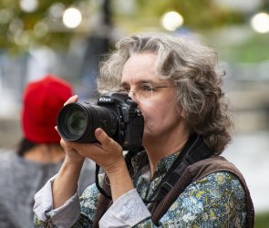 Person mit grauen lockigen Haaren und Brille hält eine Kamera zum Fotografieren vor das Gesicht