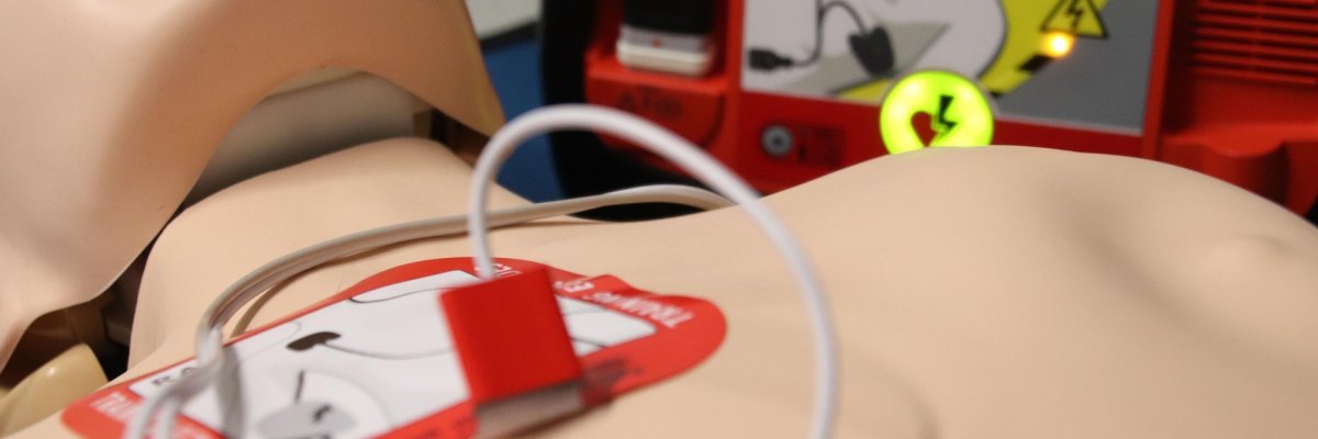 Dummy liegt mit Elektrodenpads auf der Brust vor einem geöffneten Defibrillator mit beleuchteter Anleitung