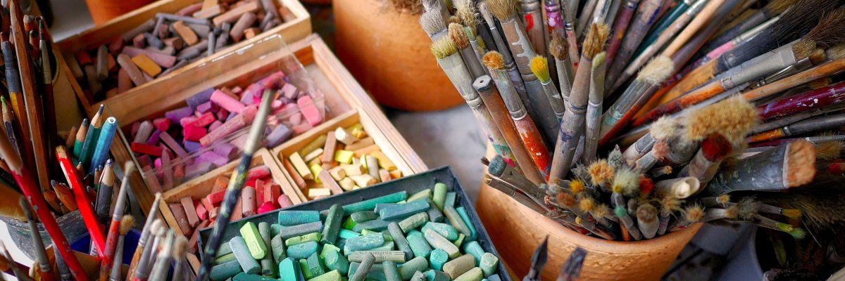 Mehrere Töpfe mit unterschiedlich großen und benutzten Pinseln sowie Schachteln mit bunten Pastellkreiden auf einem Tisch