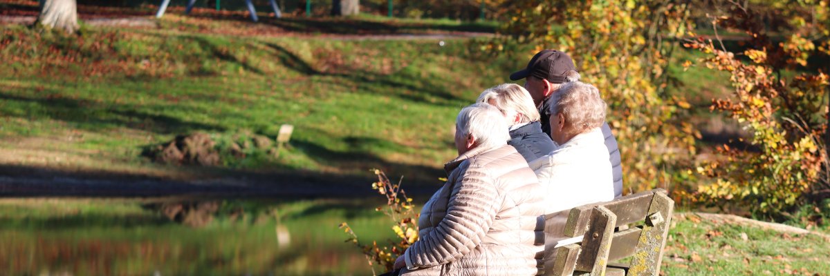 Senioren Drei ältere Personen sitzen auf einer Parkbank am Ufer eines Teichs im herbstlichen Park