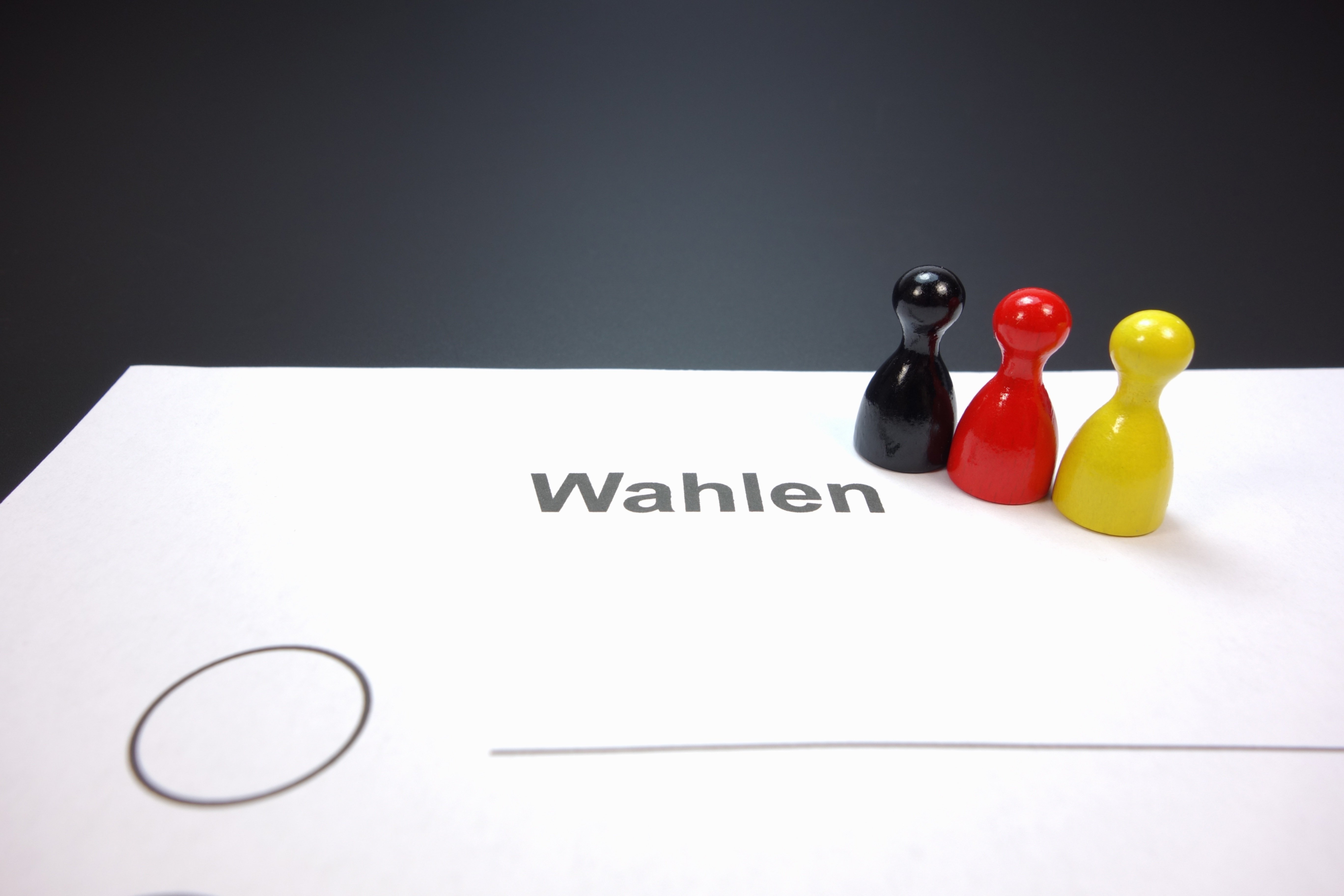 Drei Spielfiguren in den Farben Schwarz, Rot und Gelb stehen auf einem weißen Blatt mit dem Wort 'Wahlen' und einem leeren Auswahlkreis