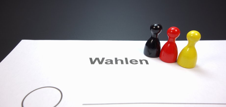 Wahlen Drei Spielfiguren in den Farben Schwarz, Rot und Gelb stehen auf einem weißen Blatt mit dem Wort 'Wahlen' und einem leeren Auswahlkreis