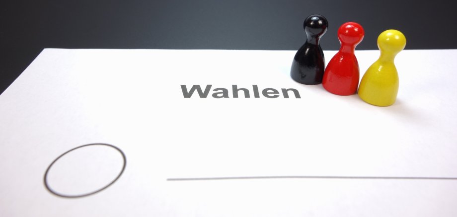 Drei Spielfiguren in den Farben Schwarz, Rot und Gelb stehen auf einem weißen Blatt mit dem Wort 'Wahlen' und einem leeren Auswahlkreis