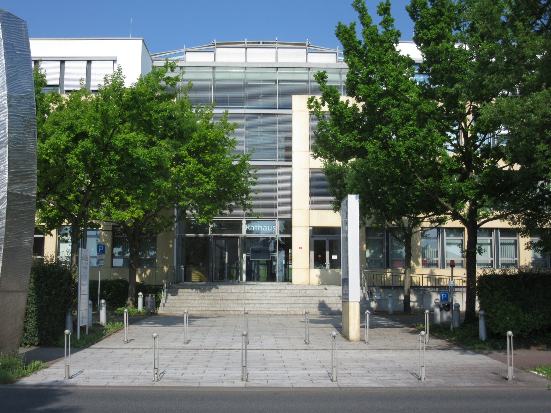 Eingang vom Rathaus Bad Vilbel, ein modernes Gebäude mit Glasfassade und Treppe, umgeben von Bäumen und mehreren Parkplätzen