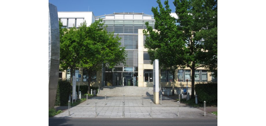 Eingang vom Rathaus Bad Vilbel, ein modernes Gebäude mit Glasfassade und Treppe, umgeben von Bäumen und mehreren Parkplätzen