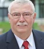 Älterer Mann (Ehrenstadtverordnetenvorsteher Herbert Anders) mit grauen Haaren, Brille, Schnurrbart, dunkelblauem Anzug, weißem Hemd und roter Krawatte vor unscharfem Hintergrund