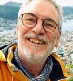 Älterer Mann (Ehrenstadtverordneter Klaus Arabin) mit Brille, grauem Bart und gelber Jacke lächelt vor einer Stadt- und Berglandschaft