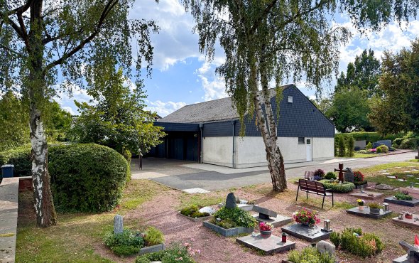 Friedhof mit Gräbern, Bänken und einem schlichten Gebäude im Hintergrund unter blauem Himmel mit Wolken