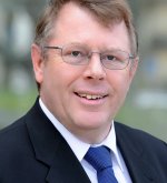 Porträt eines lächelnden Mannes (Ehrenstadtverordneter Edwin Lotz) mit Brille, Anzug und blauer Krawatte vor unscharfem Hintergrund