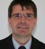 Porträt eines Mannes mit kurzen braunen Haaren, Brille, Hemd und dunkler Uniform vor weißem Hintergrund