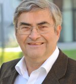 Älterer Mann (Ehrenstadtrat Klaus Minkel) mit grauen Haaren, Brille und dunklem Sakko lächelt in die Kamera vor unscharfem Hintergrund mit Gebäuden