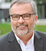 Mann mit Brille (Ehrenortsvorsteher Karl Peter Schäfer), grauem Haar und Bart trägt weißen Hemdkragen unter dunklem Sakko und lächelt in die Kamera