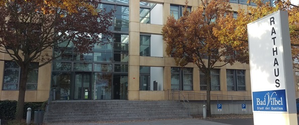 Rathaus Eingang eines modernen Gebäudes mit großen Fenstern und herbstlich gefärbten Bäumen davor, rechts ein Schild mit der Aufschrift 'Rathaus Bad Vilbel Stadt der Quellen'