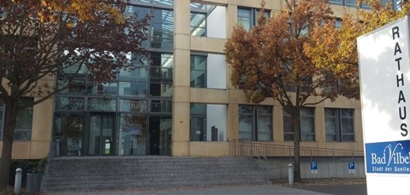 Rathaus Eingang eines modernen Gebäudes mit großen Fenstern und herbstlich gefärbten Bäumen davor, rechts ein Schild mit der Aufschrift 'Rathaus Bad Vilbel Stadt der Quellen'