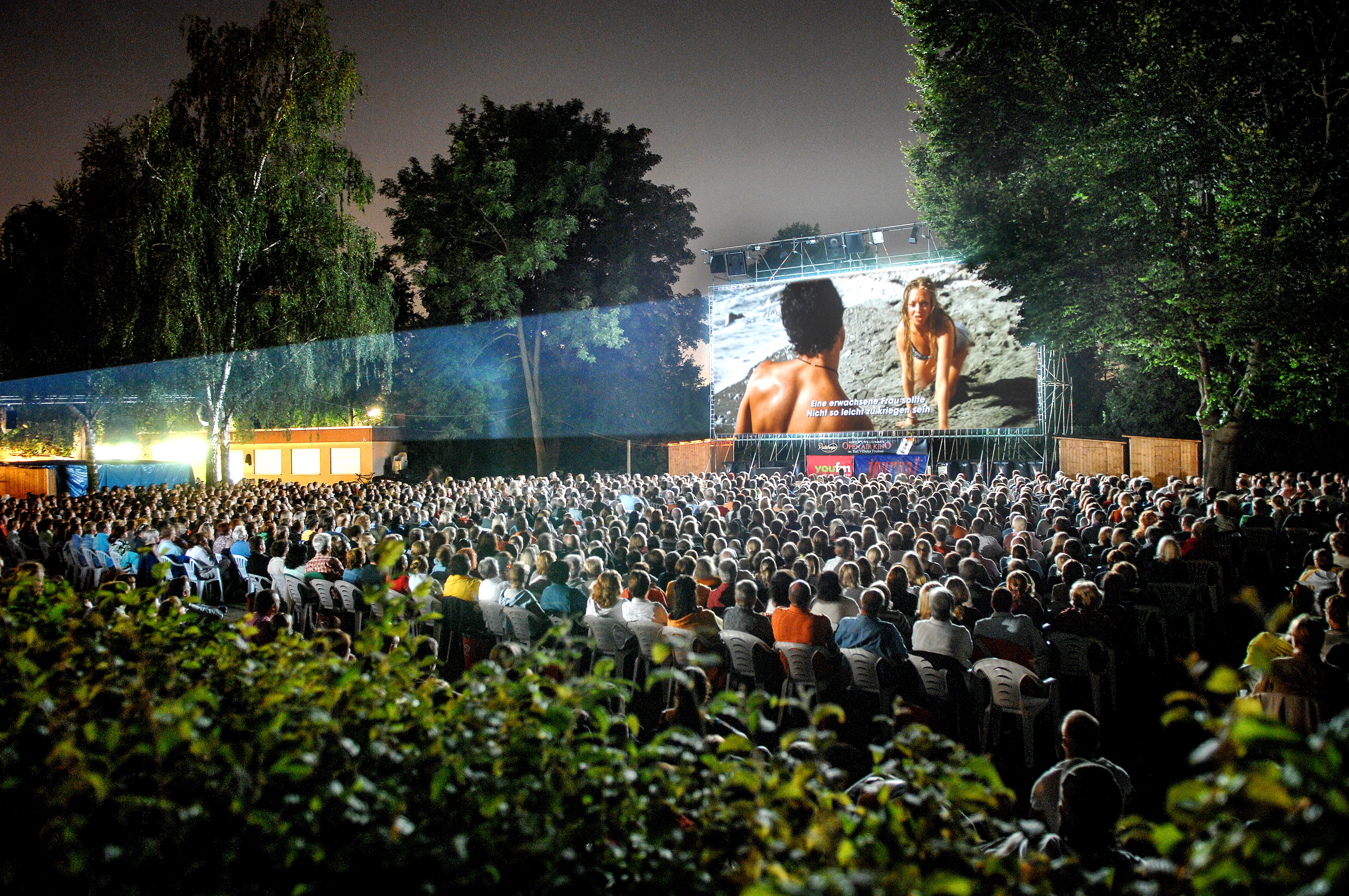 Open-Air-Kino Bad Vilbel Große Menschenmenge sitzt bei Nacht im Open-Air-Kino Bad Vilbel vor einer großen Leinwand, auf der ein Film mit zwei Personen gezeigt wird, umgeben von Bäumen und Hecken.