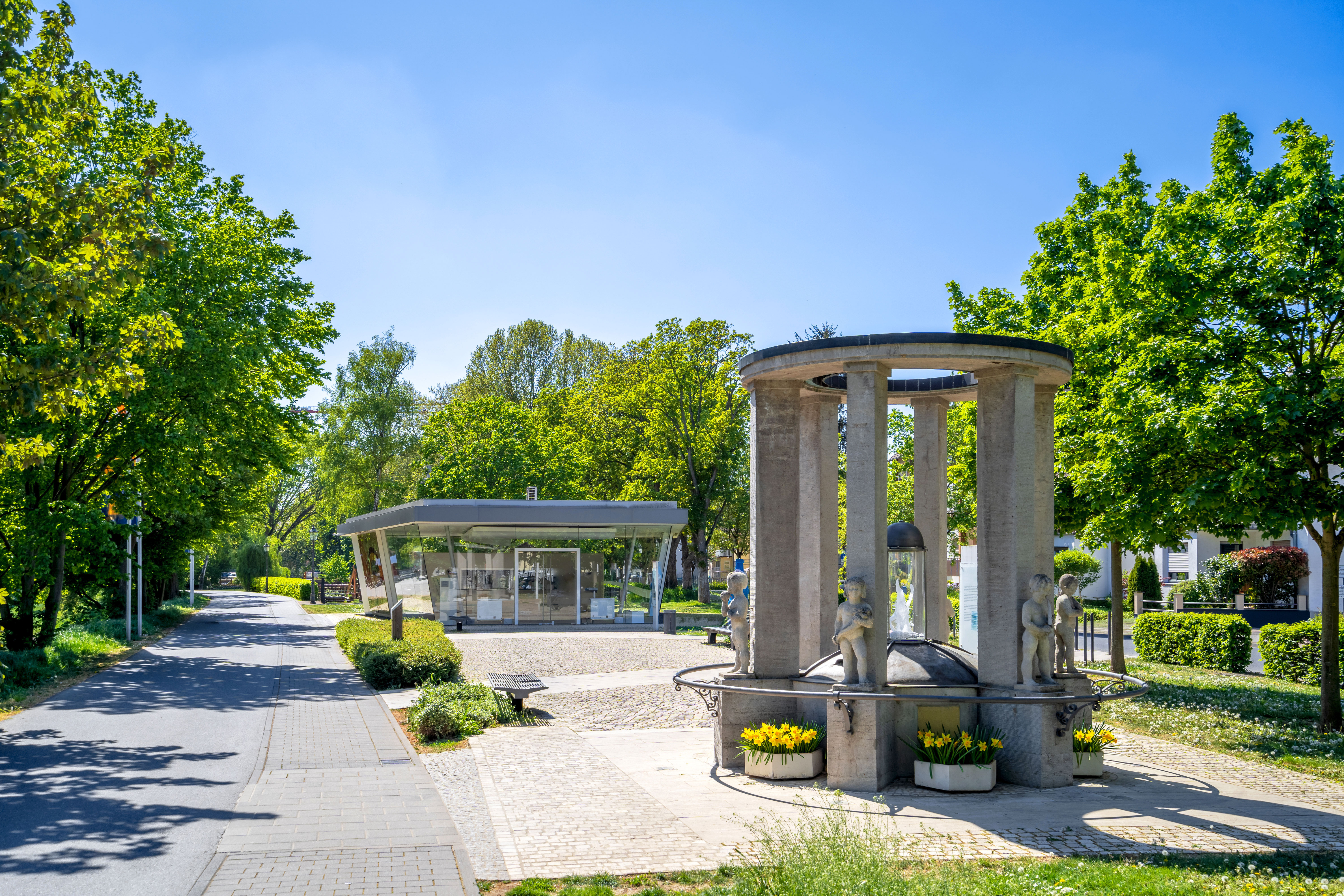 Runder Brunnen mit Säulen und Blumenkästen neben einem modernen Glasgebäude an einem von Bäumen gesäumten Weg
