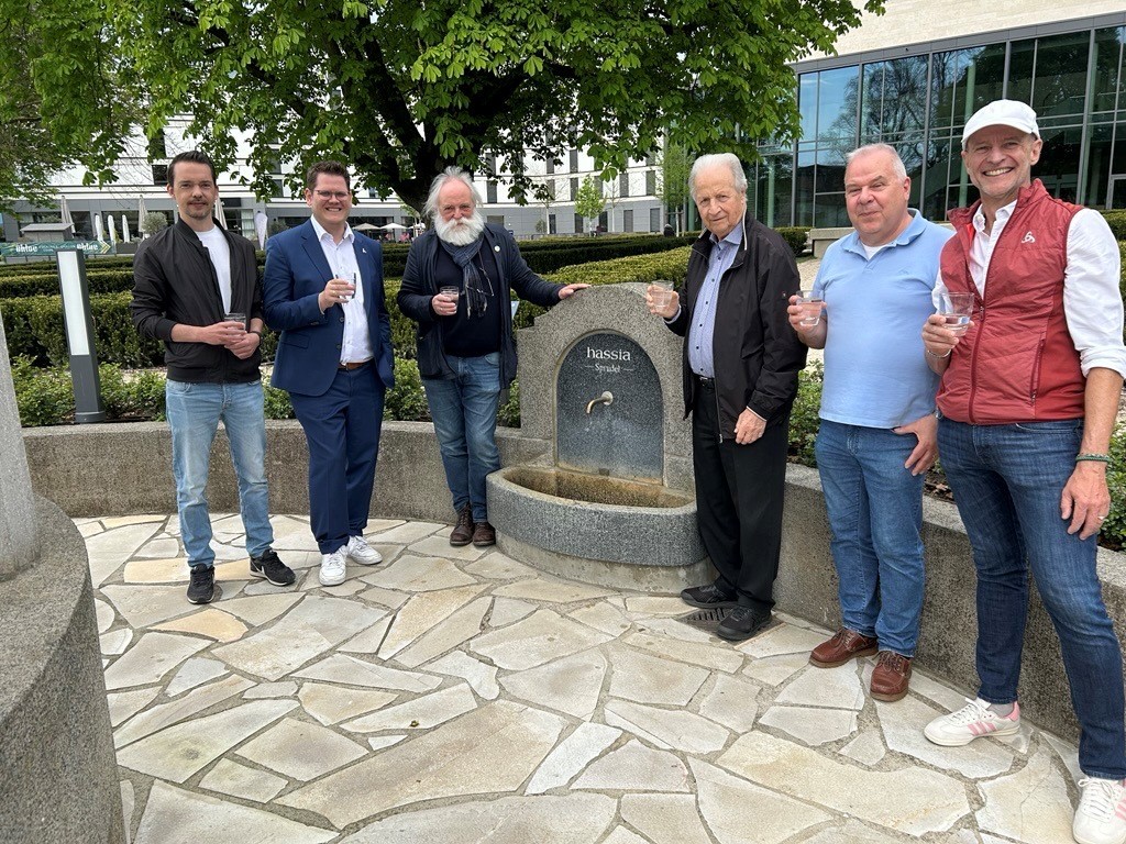 Sechs Personen stehen um einen runden Trinkbrunnen aus Stein mit der Aufschrift 'Hassia Quelle' und halten Gläser in der Hand