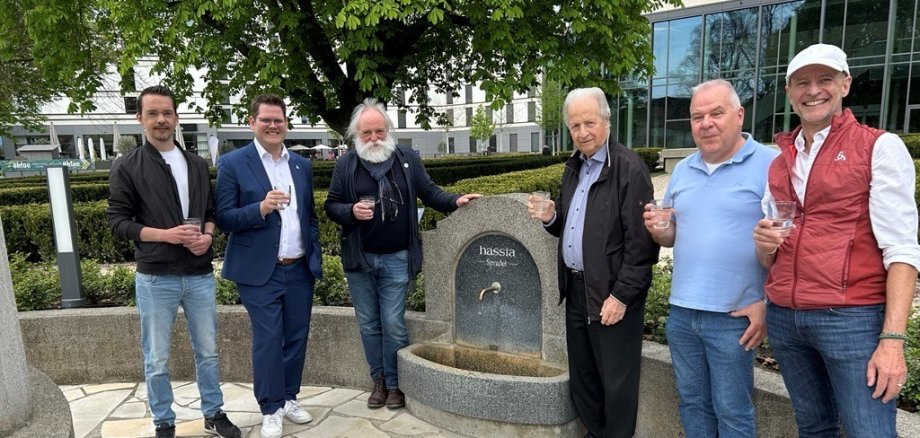 Sechs Personen stehen um einen runden Trinkbrunnen aus Stein mit der Aufschrift 'Hassia Quelle' und halten Gläser in der Hand