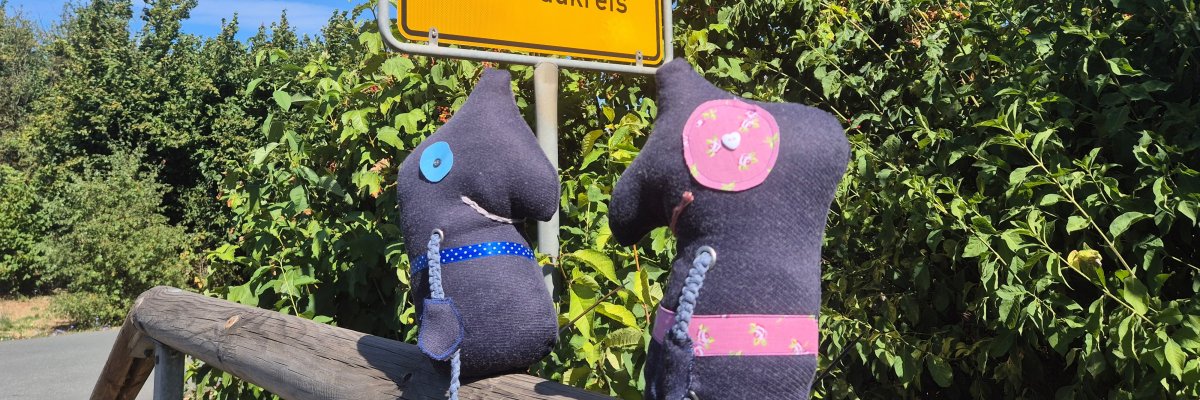 Europäische Mobilitätswoche Zwei dunkelblaue Stofffiguren mit bunten Applikationen sitzen auf einem Holzgeländer vor einem gelben Ortsschild mit der Aufschrift 'Bad Vilbel Wetteraukreis'