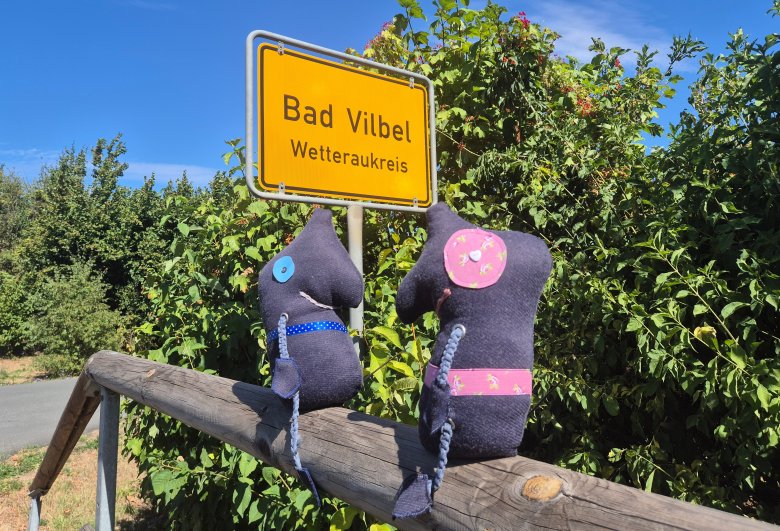 Europäische Mobilitätswoche Zwei dunkelblaue Stofffiguren mit bunten Applikationen sitzen auf einem Holzgeländer vor einem gelben Ortsschild mit der Aufschrift 'Bad Vilbel Wetteraukreis'