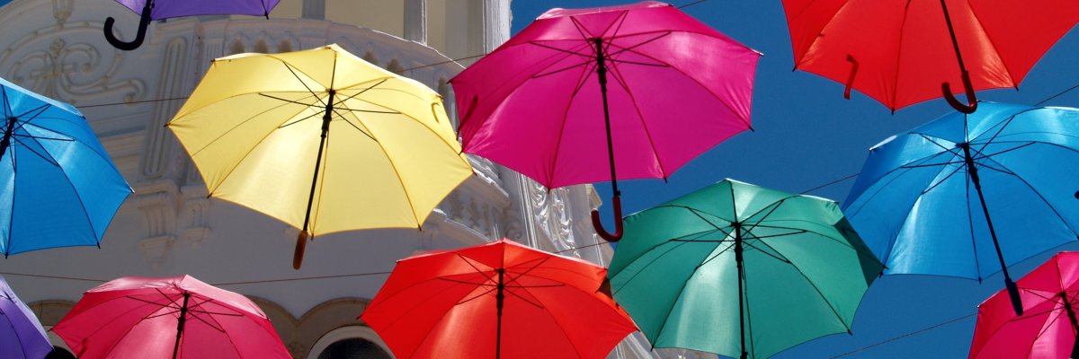 Bunte Regenschirme hängen über einer Straße vor blauem Himmel und einem weißen Gebäude