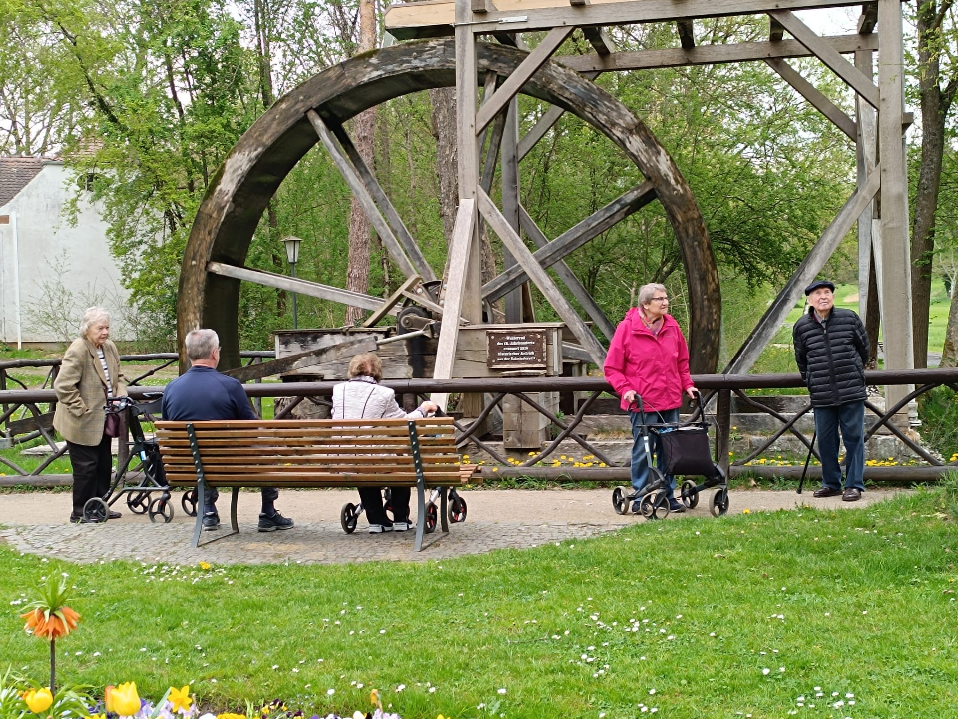 Fahrten für mobilitätseingeschränkte Personen Mehrere ältere Menschen mit Rollatoren sitzen und stehen vor einem großen hölzernen Wasserrad in einem Park