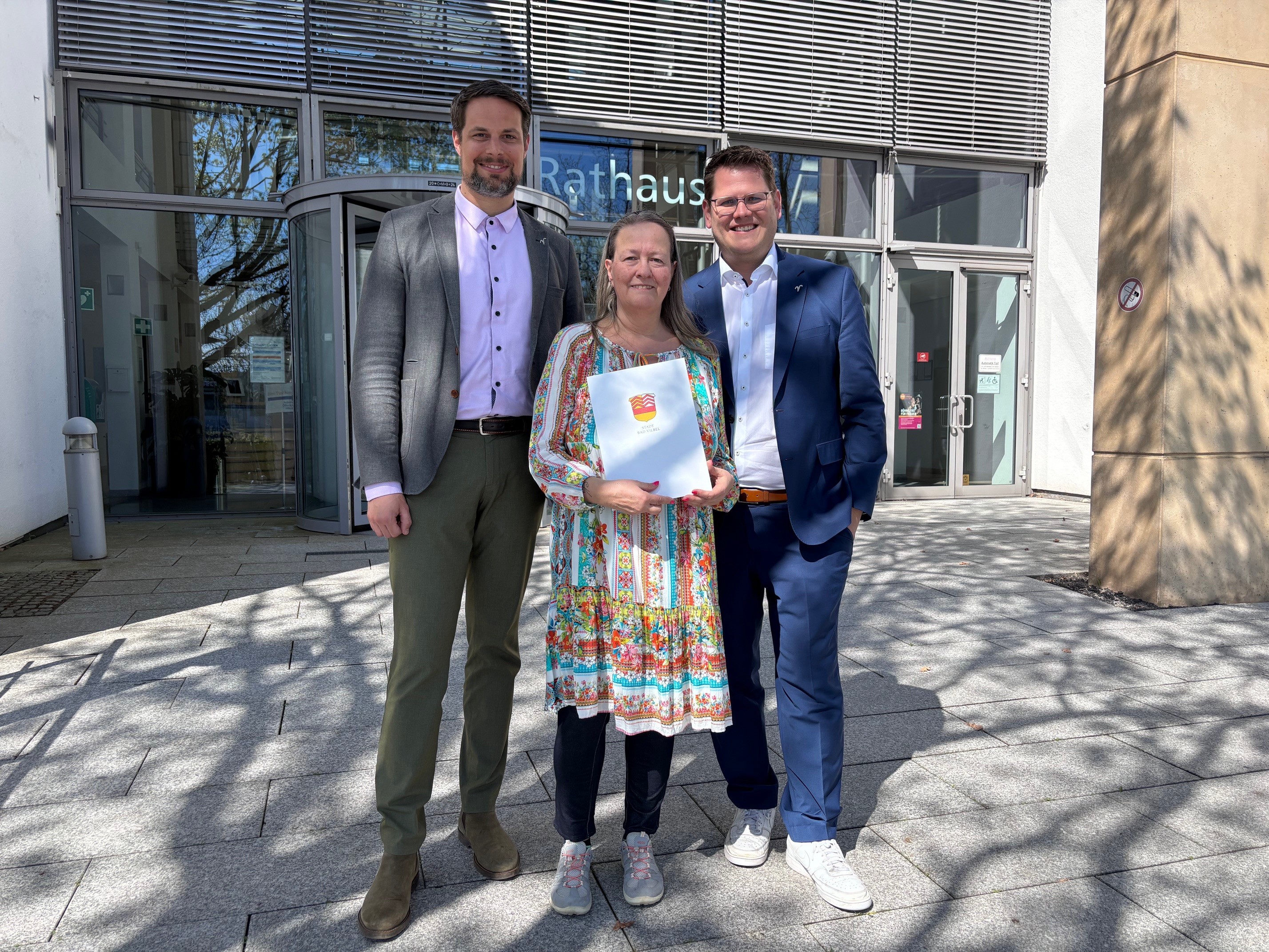 Drei Personen stehen vor dem Rathaus Bad Vilbel, die mittlere Person, Frau Elke Bär, hält eine Mappe mit einem bunten Wappen