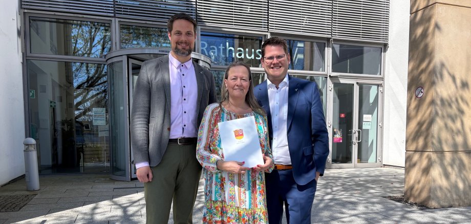 Drei Personen stehen vor dem Rathaus Bad Vilbel, die mittlere Person, Frau Elke Bär, hält eine Mappe mit einem bunten Wappen
