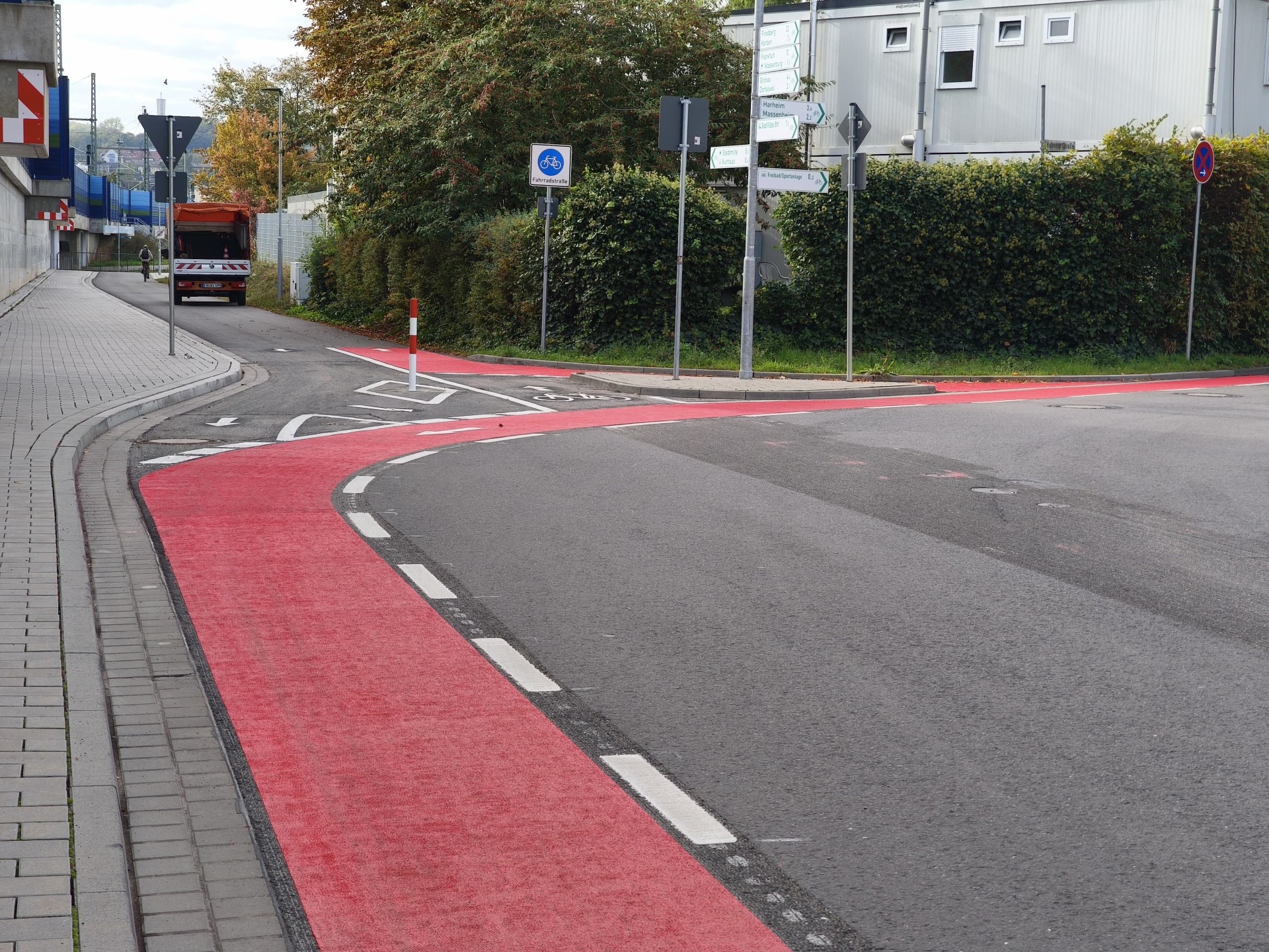 Straße mit rotem Fahrradstreifen, Gehweg mit Bordstein und mehreren Verkehrsschildern neben einer Hecke und einem Gebäude