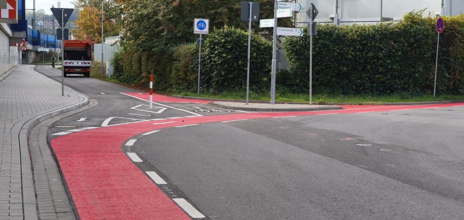 Straße mit rotem Fahrradstreifen, Gehweg mit Bordstein und mehreren Verkehrsschildern neben einer Hecke und einem Gebäude